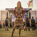 A woman modeling a hooded longline cloak with an intricate, colorful abstract print featuring gold, orange, red, and purple tones, at a music festival or outdoor concert